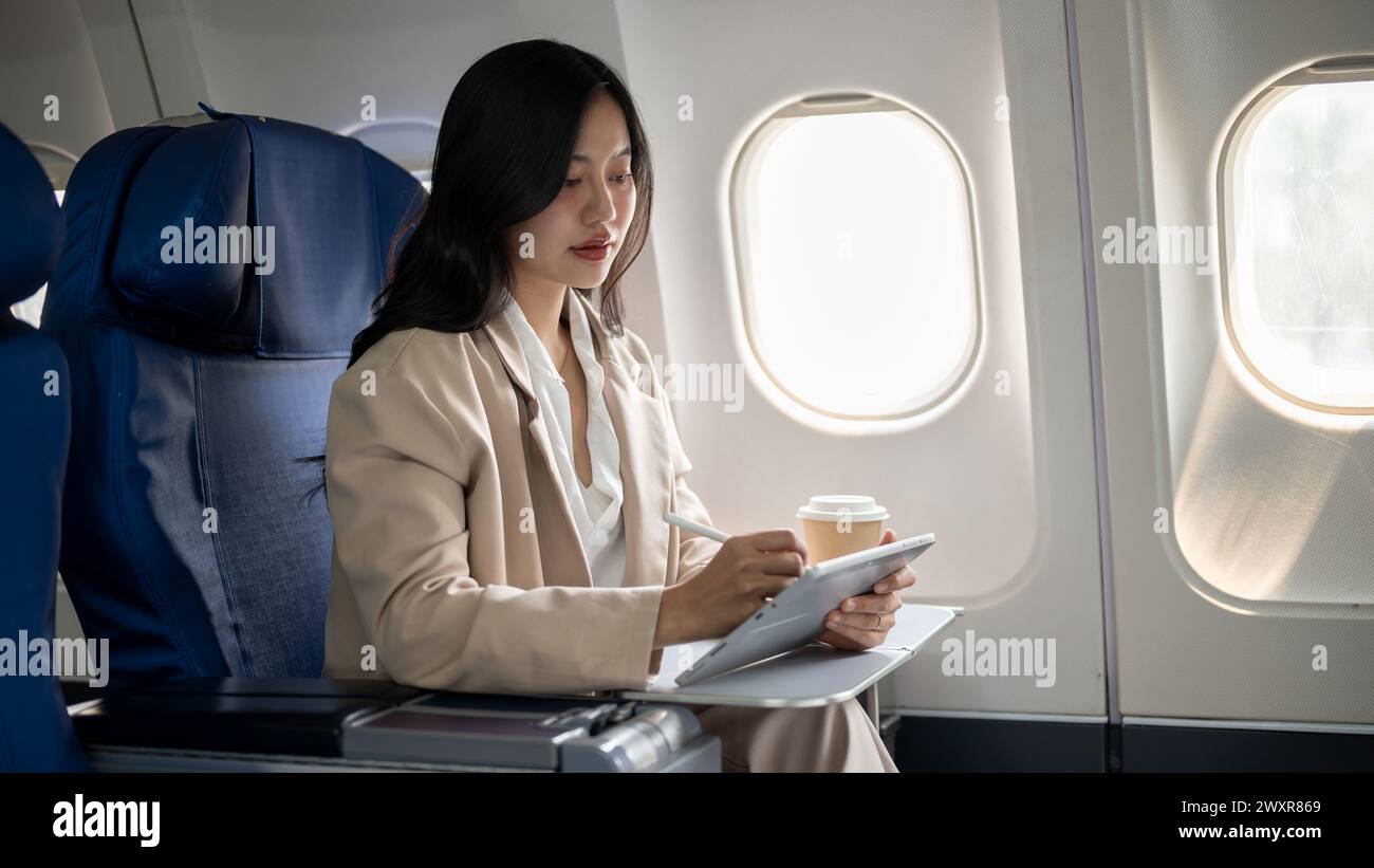 Une femme d'affaires asiatique confiante et travailleuse en costume d'affaires se concentre sur son travail sur sa tablette numérique alors qu'elle s'envole vers une autre ville pour un busi Banque D'Images