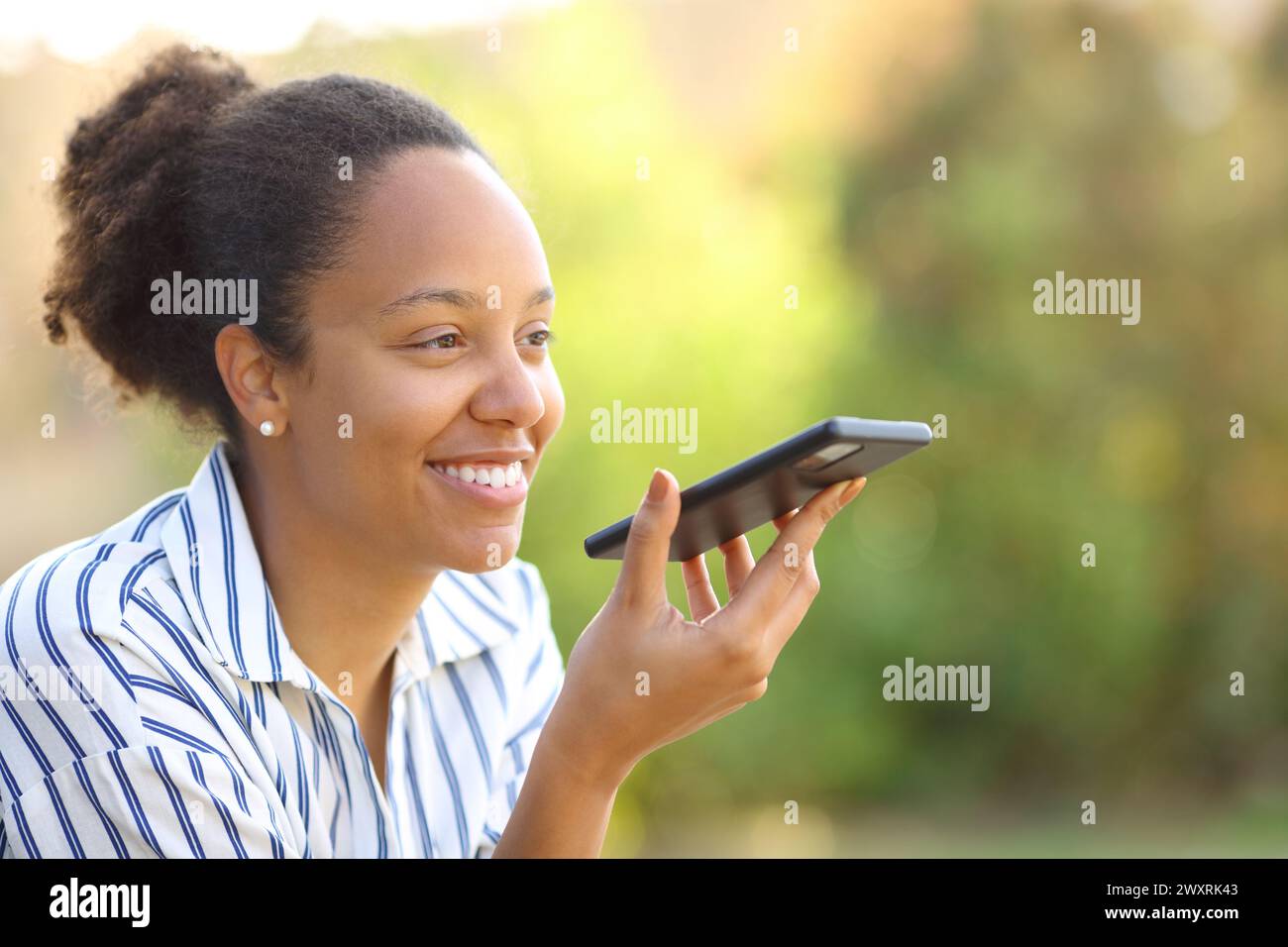 Femme noire heureuse dictant un message sur un téléphone portable dans un parc Banque D'Images