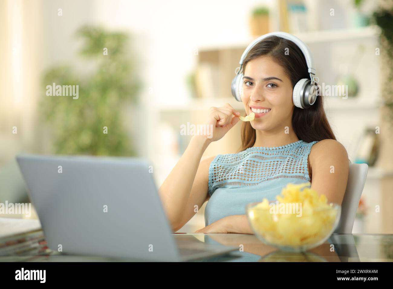 Femme avec ordinateur portable et casque mangeant des chips regardant la caméra à la maison Banque D'Images