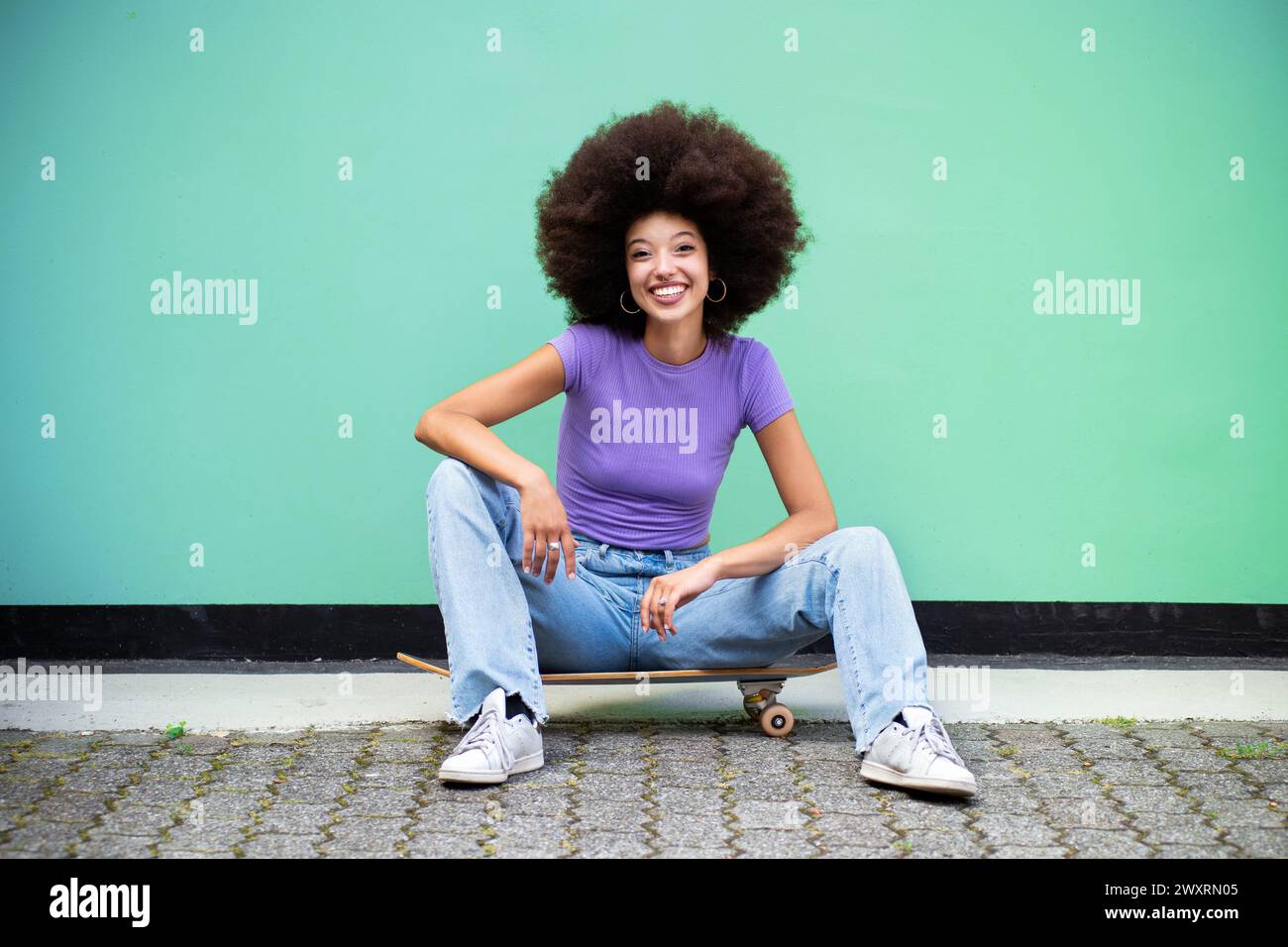 Corps complet de jeune femme marocaine joyeuse avec coiffure afro et dans des vêtements décontractés regardant la caméra tout en étant assis à la lumière du jour sur le skateboard sur la str Banque D'Images