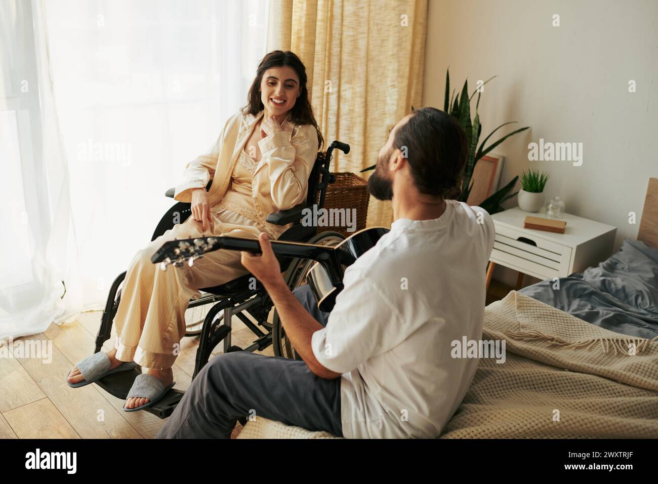 femme joyeuse handicapée en fauteuil roulant regardant son mari barbu jouer de la guitare pour elle Banque D'Images