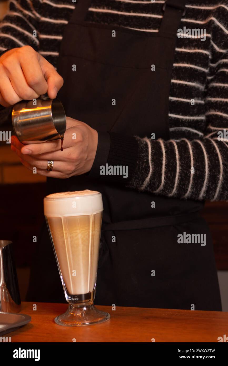 Un barista habillé d'une élégante chemise rayée et d'un tablier noir verse du sirop dans un verre de café aromatique sur une table en bois dans un éclairage doux, créant ainsi Banque D'Images