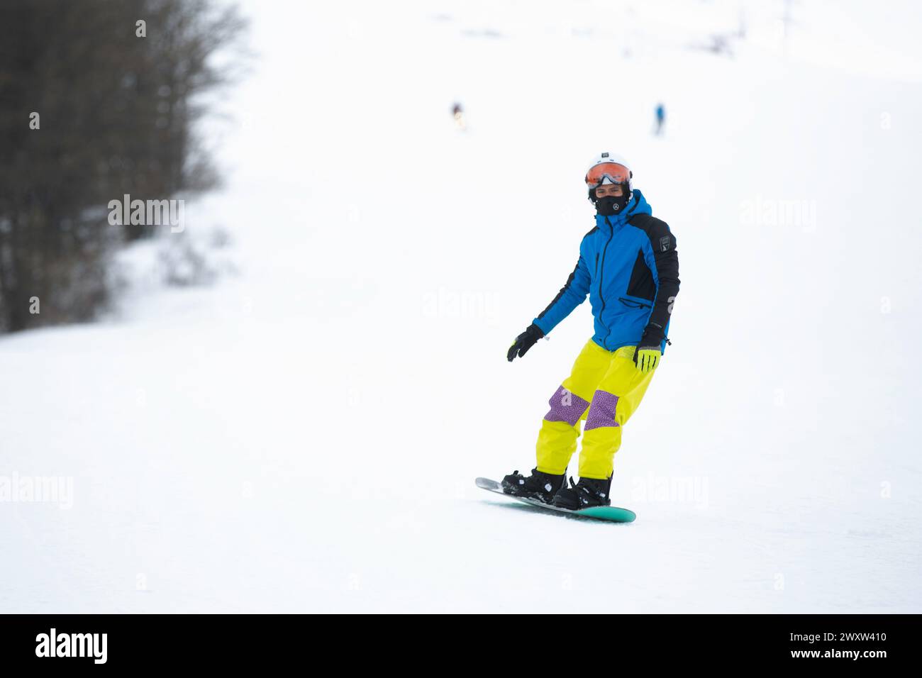 Un snowboarder masculin dans une veste bleue et un pantalon jaune sculptant une montagne enneigée Banque D'Images