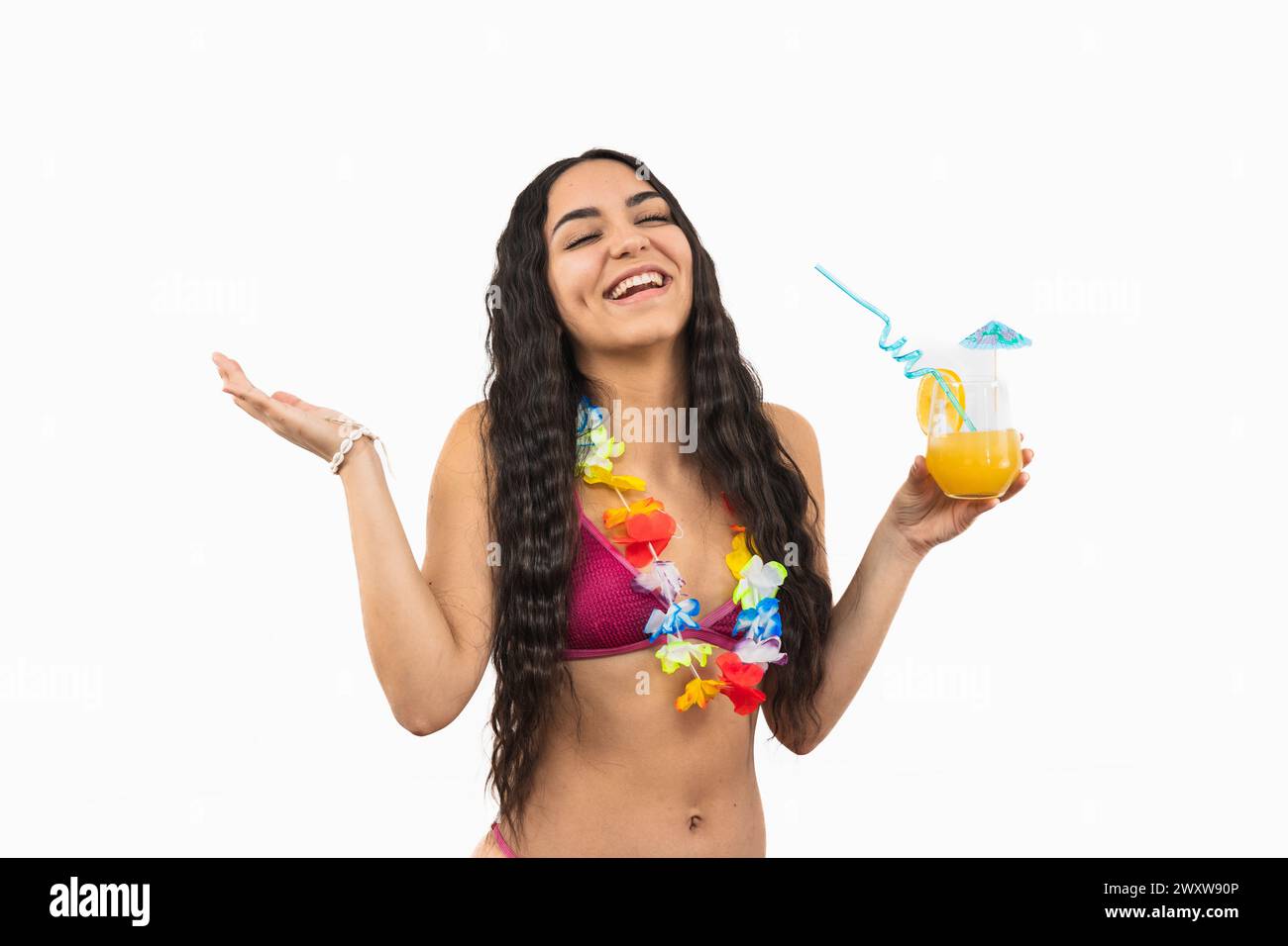 Joyeuse jeune femme latina portant un bikini rit joyeusement tout en tenant un cocktail d'été, orné d'un collier de fleurs autour du cou, sur un soleil Banque D'Images