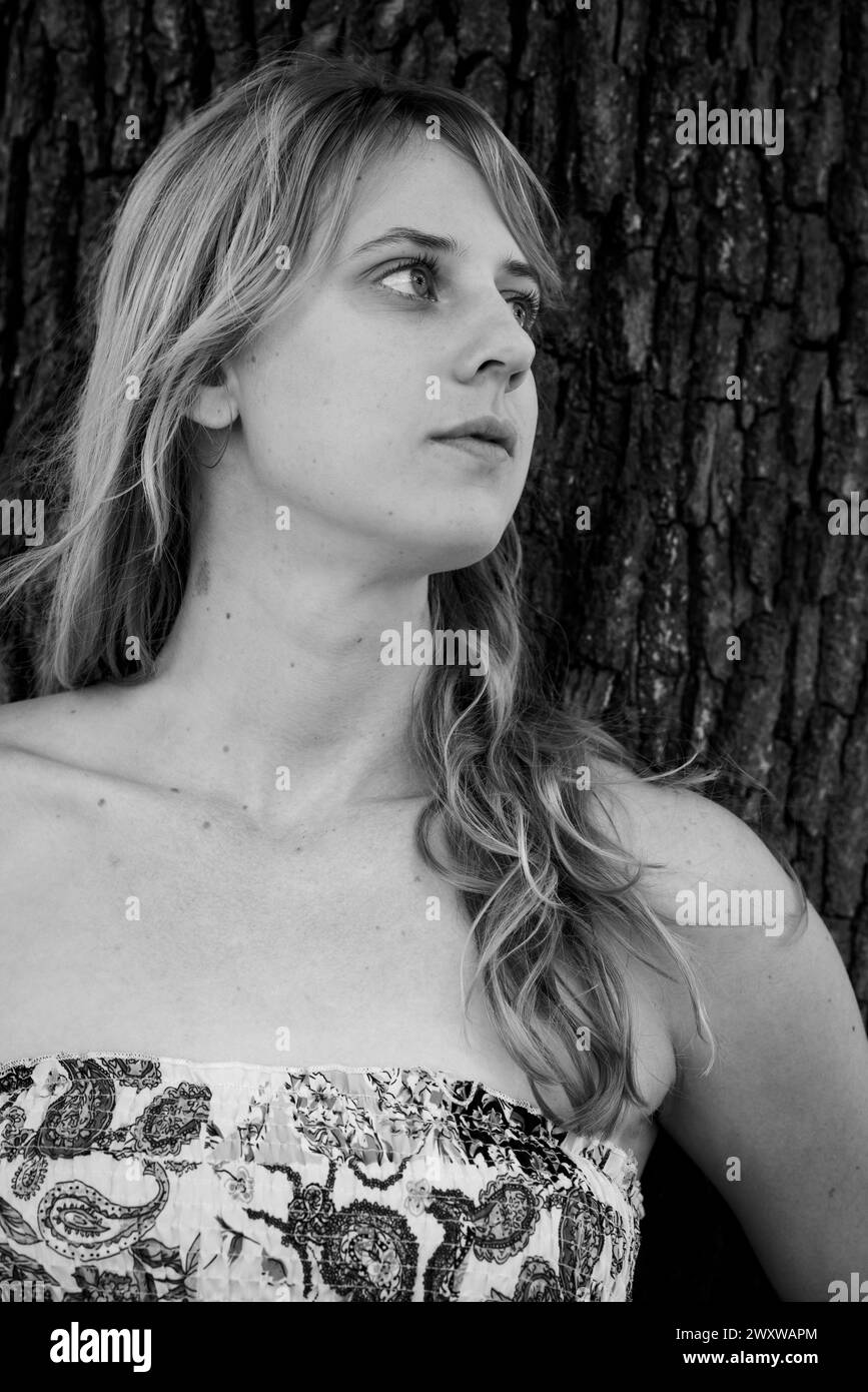 Une photo en niveaux de gris d'une fille blonde appuyée contre un arbre Banque D'Images