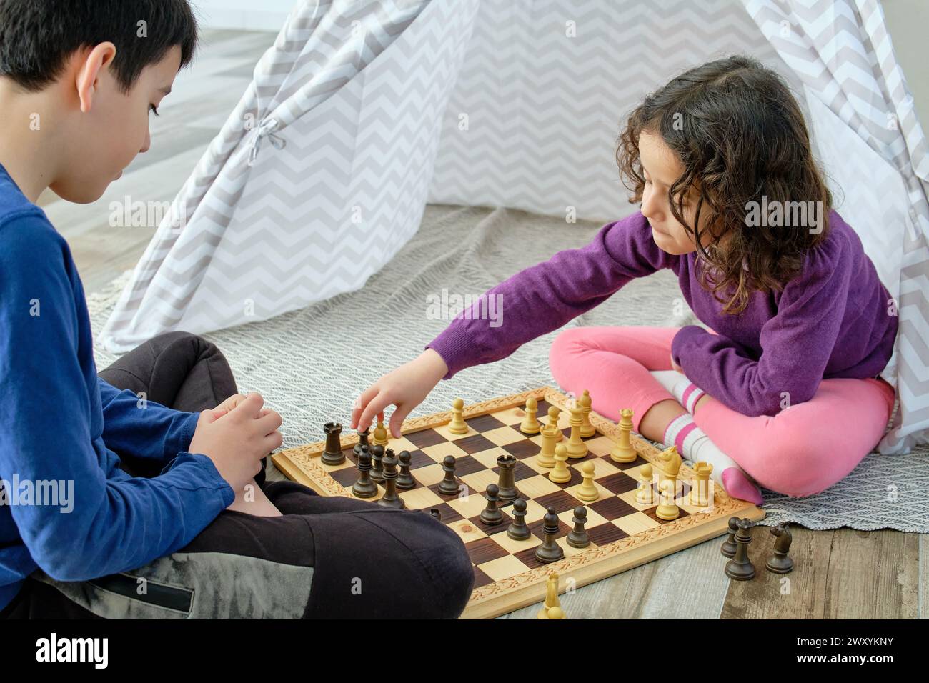 Deux enfants, un garçon et une fille, contemplent soigneusement leur prochain mouvement tout en jouant aux échecs sur une planche de bois assise sur le sol Banque D'Images