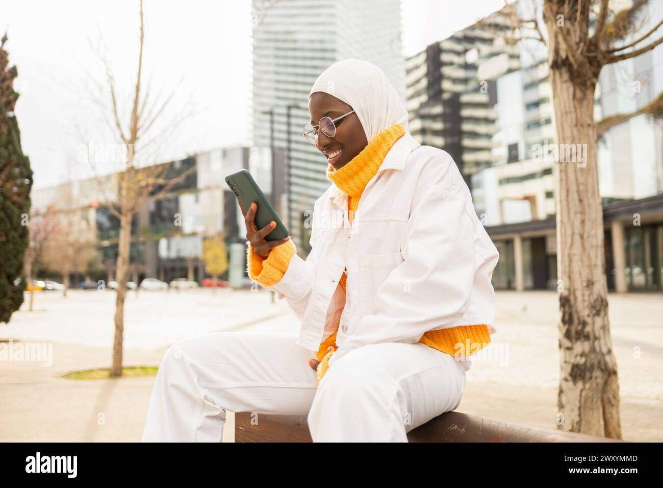 Femme afro-américaine élégante en tenue blanche en utilisant son téléphone tout en étant assis dans un environnement de ville moderne. Banque D'Images