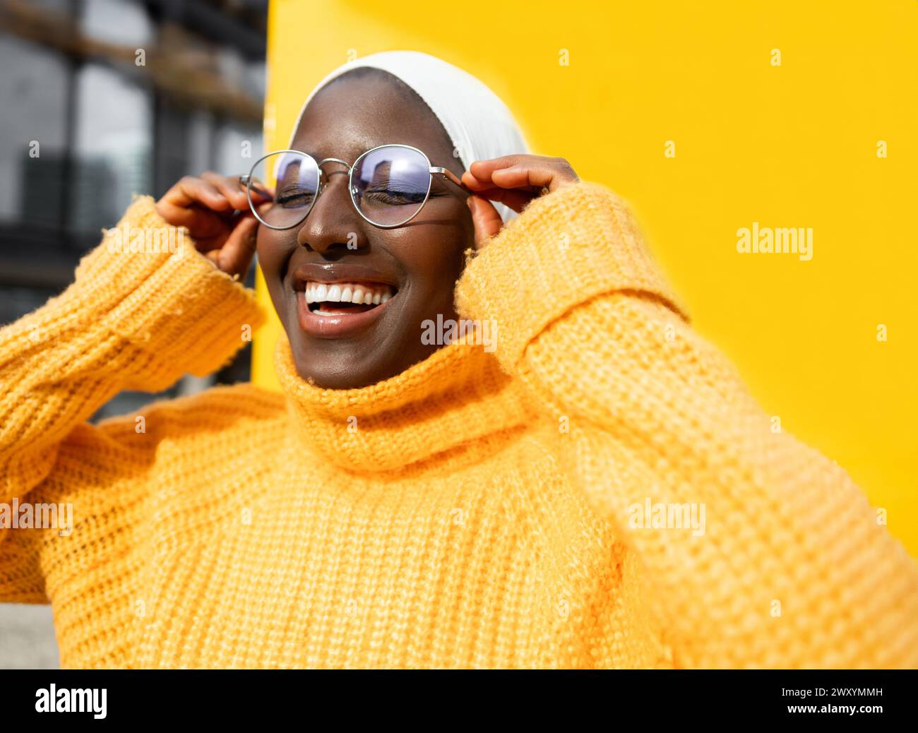 Femme noire souriante en pull jaune ajustant les lunettes sur un fond jaune vif, regardant la caméra. Banque D'Images