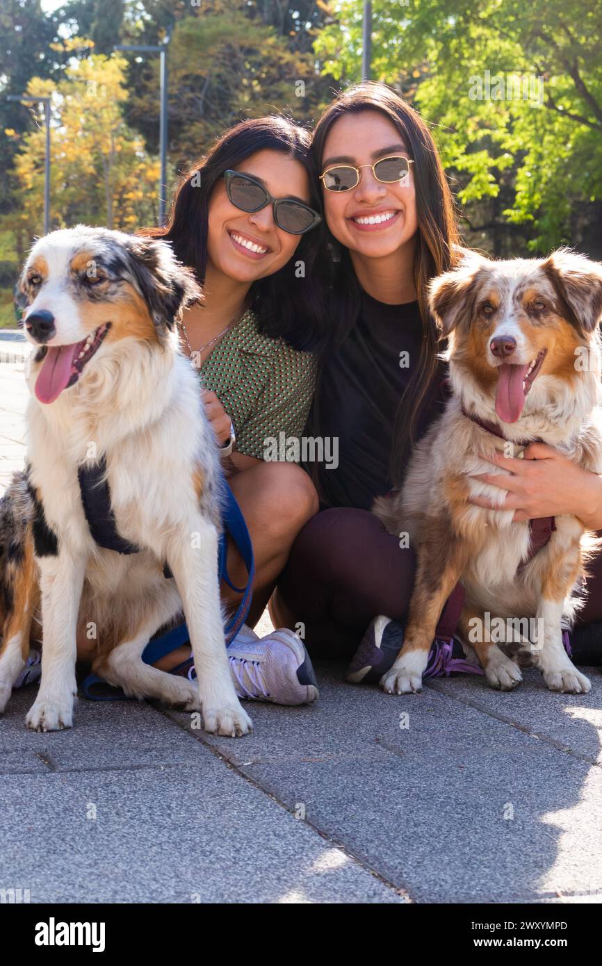Deux jeunes femmes heureuses s'agenouillant et posant avec leurs chiens bergers australiens joyeux dans un parc ensoleillé Banque D'Images