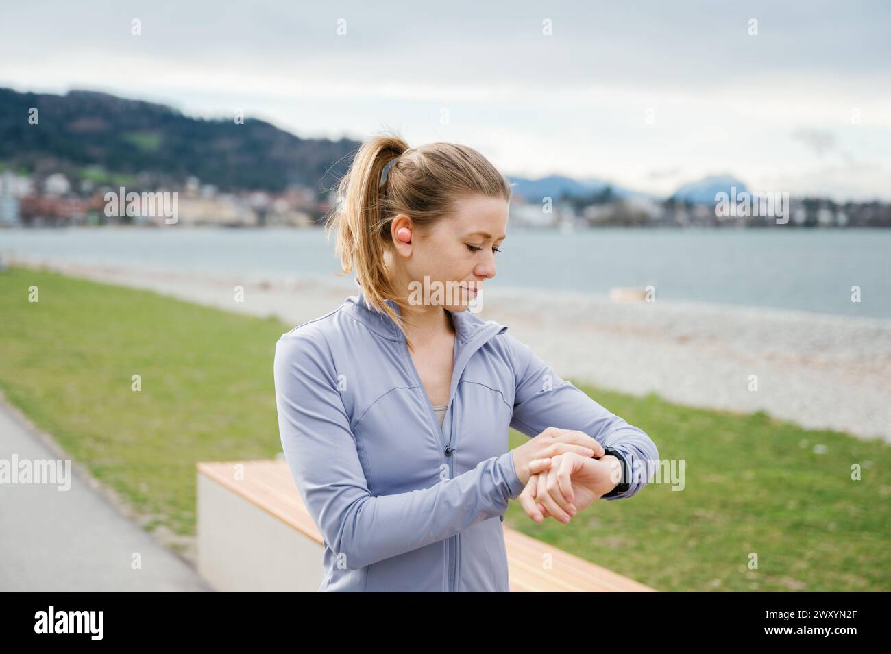 Une femme concentrée en vêtements sportifs utilise sa smartwatch pour surveiller son entraînement au bord du lac, avec des paysages naturels autour Banque D'Images