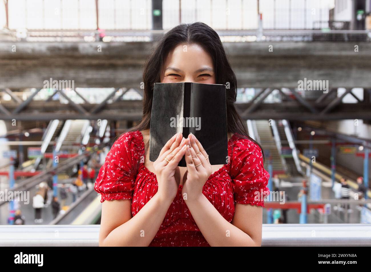 Une femme joyeuse se tient dans une gare très fréquentée, cachant ludique son visage derrière un livre noir, vêtue d'une robe rouge Banque D'Images