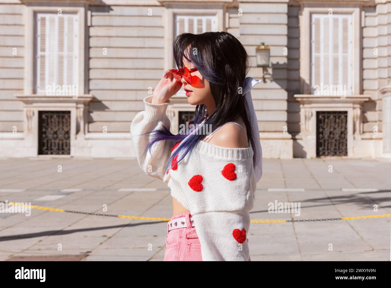 Une jeune chinoise de génération Z à la mode avec des cheveux pourpres ajuste des lunettes de soleil en forme d'étoile tout en portant un pull à motif coeur et un Jean rose Banque D'Images