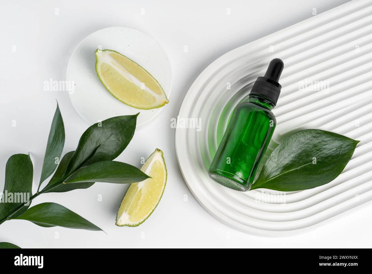 Des tranches de citron vert et des feuilles fraîches disposées artistiquement autour d'un flacon compte-gouttes vert transmettent une huile essence pure infusée d'agrumes sur un fond ondulé moderne Banque D'Images
