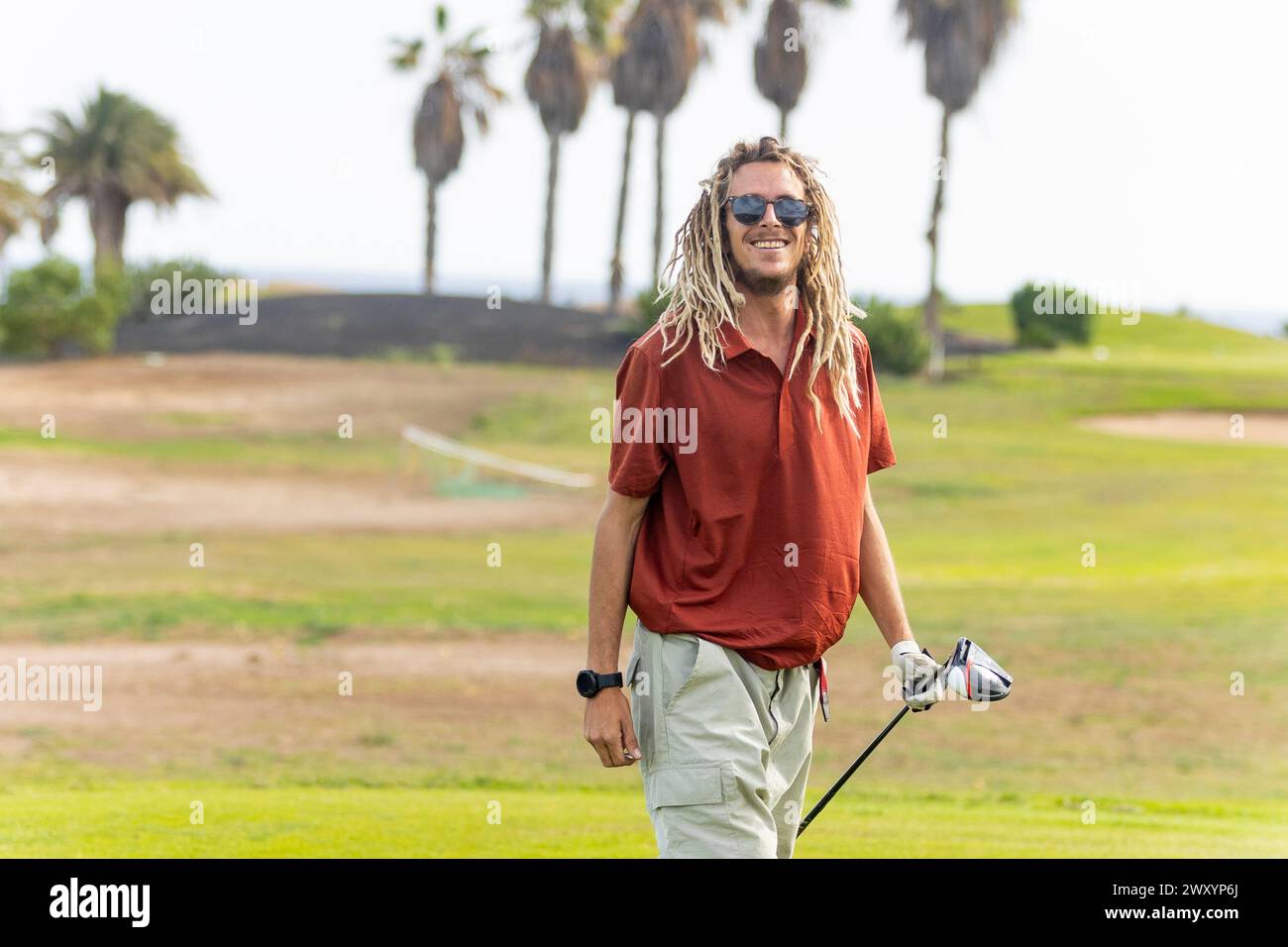 Un golfeur aux cheveux blonds détendu avec des dreadlocks sourires tout en tenant un club, prêt pour une session de golf par une journée ensoleillée au milieu des palmiers. Banque D'Images