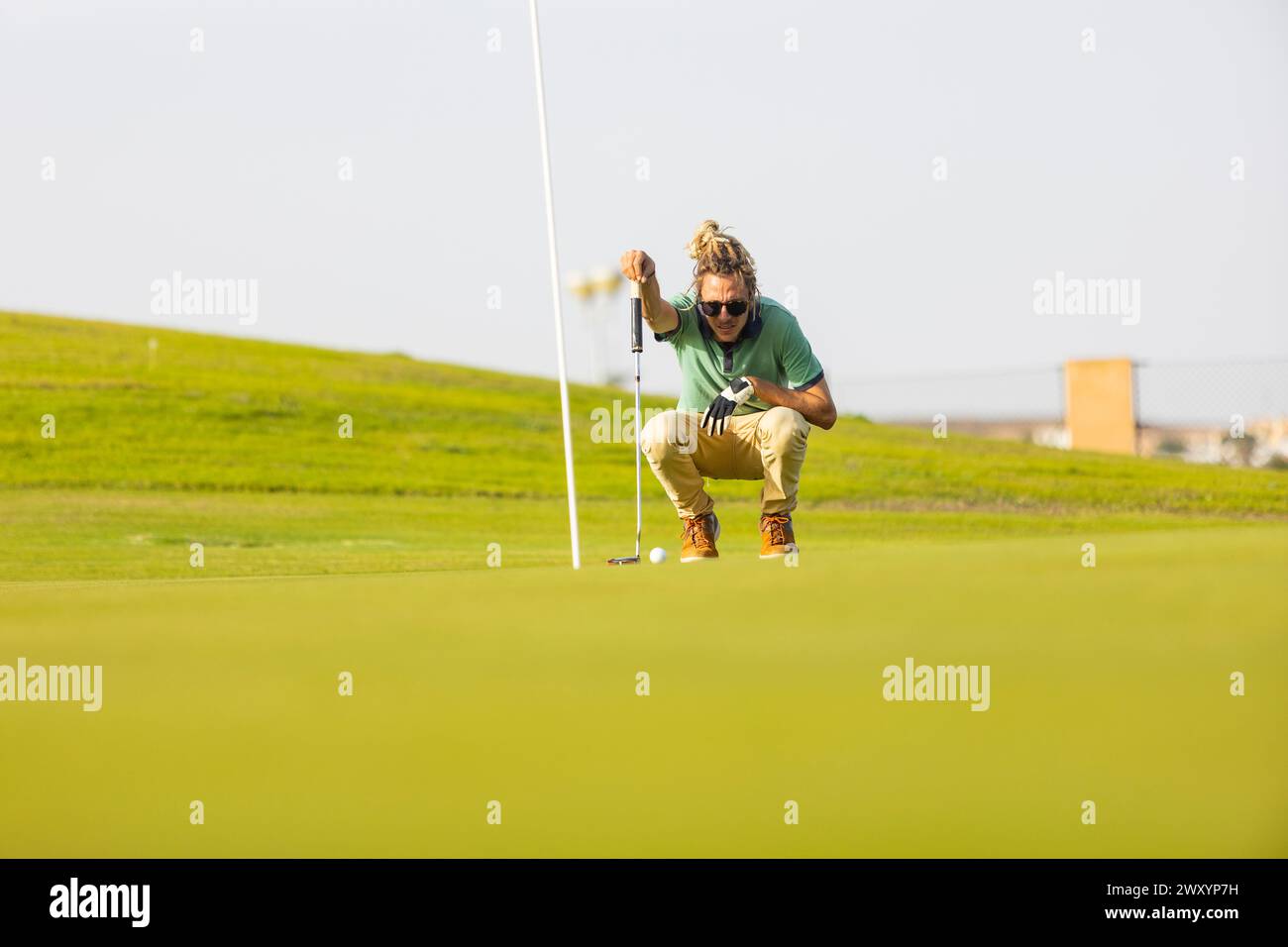 Un golfeur blond distinctif avec des dreadlocks crouches pour aligner un putt sur un terrain de golf ensoleillé, mettant en valeur un look contemporain et non conventionnel en t Banque D'Images