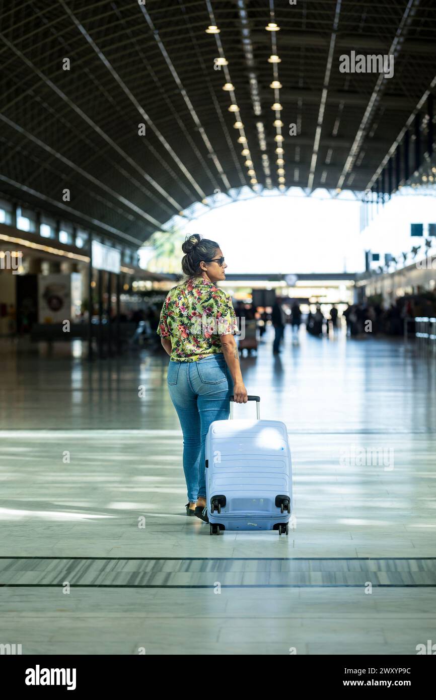 Une femme élégante marche en toute confiance dans un terminal d'aéroport moderne, tirant une valise blanche, prête pour son voyage Banque D'Images