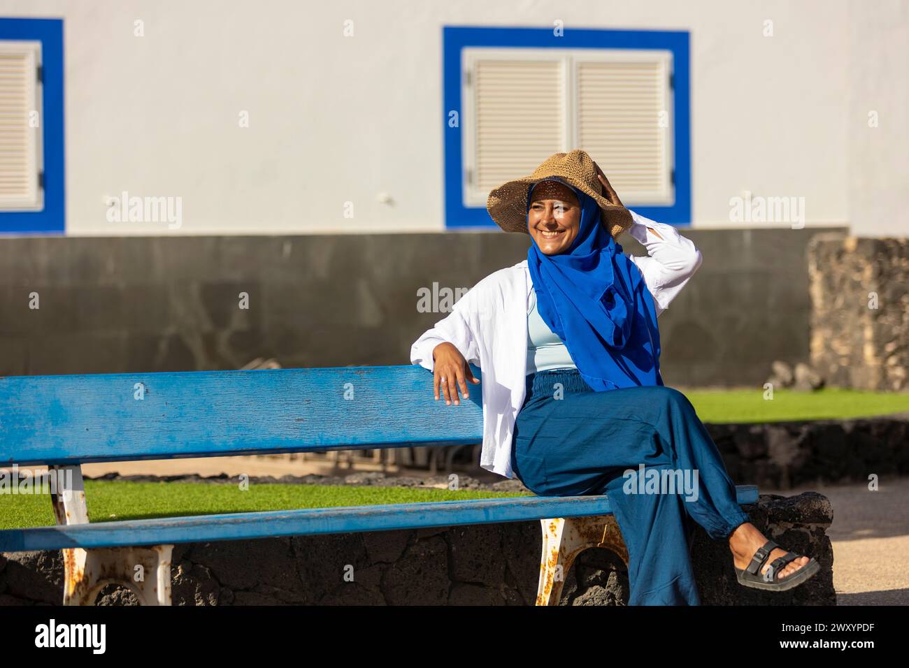 Femme multiethnique joyeuse avec un chapeau de paille et une écharpe bleue assise sur un banc bleu à l'extérieur, profitant d'une journée ensoleillée Banque D'Images