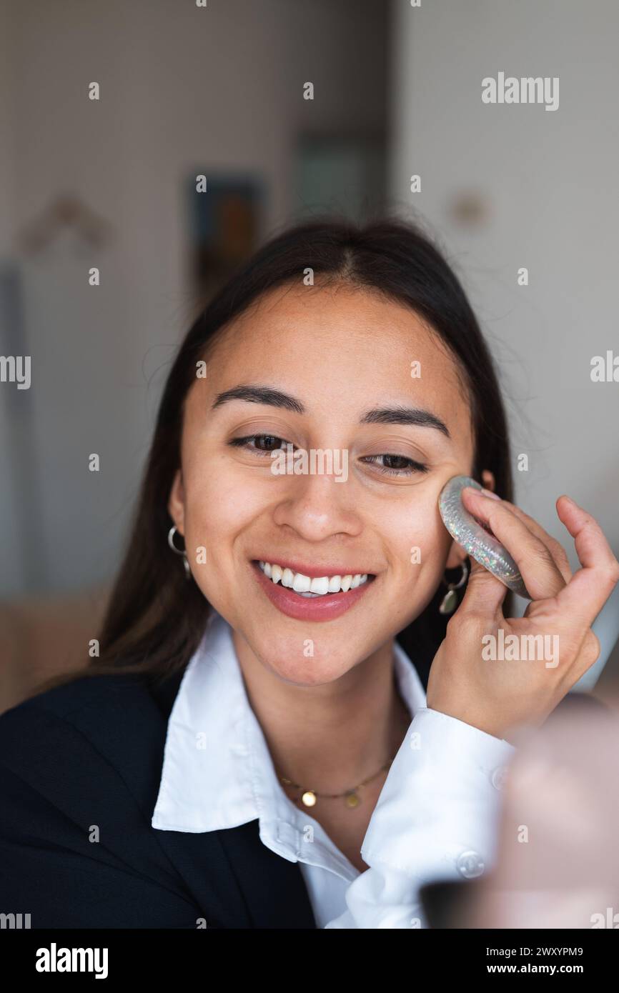 Une jeune femme joyeuse est vue appliquer le maquillage avec un pinceau, arborant une tenue professionnelle et un sourire éclatant Banque D'Images