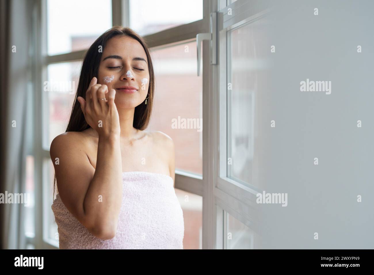 Une femme enveloppée dans une serviette appliquant un hydratant sur son visage près d'une fenêtre lumineuse, en se concentrant sur les soins de la peau Banque D'Images