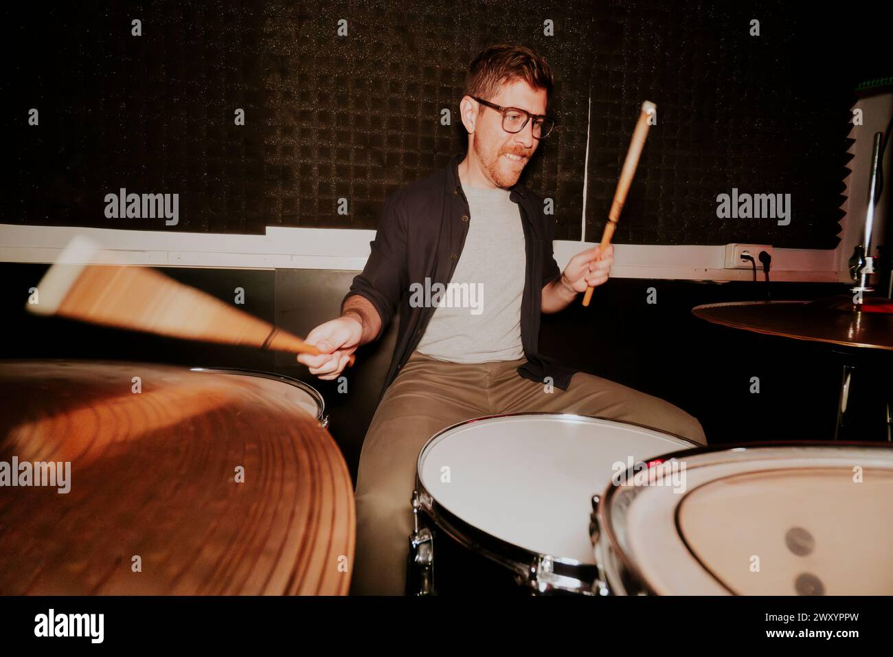 Batteur masculin énergique avec des lunettes en mouvement, jouant de la batterie avec le sourire, dans une pièce avec une ambiance sombre, ne regardant pas la caméra. Banque D'Images