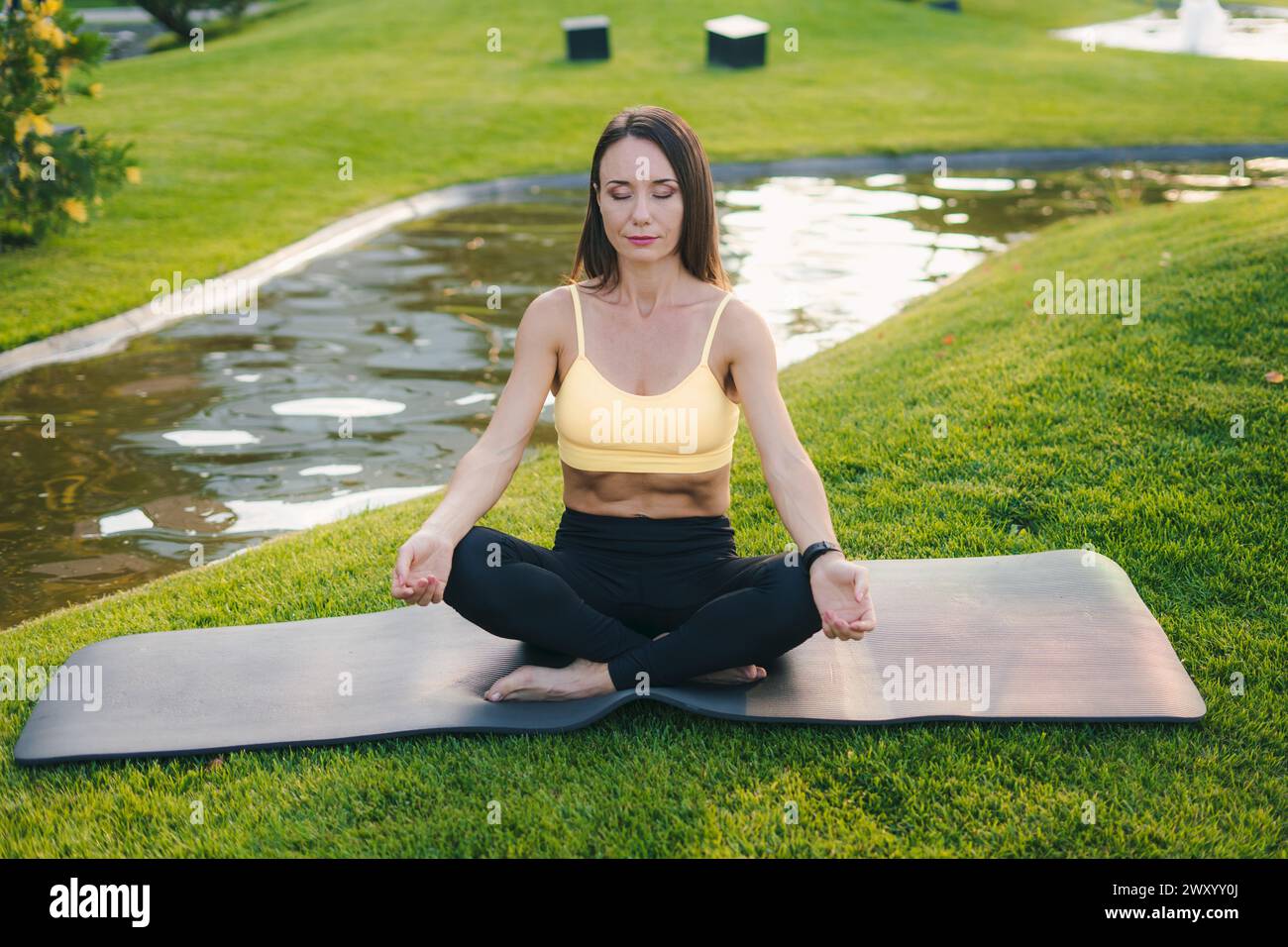 Femme brune sportive en vêtements de sport assise sur un tapis de fitness noir avec les yeux fermés dans la pose de lotus, pratiquant le yoga dans un parc public. Yoga au parc. Concept Banque D'Images