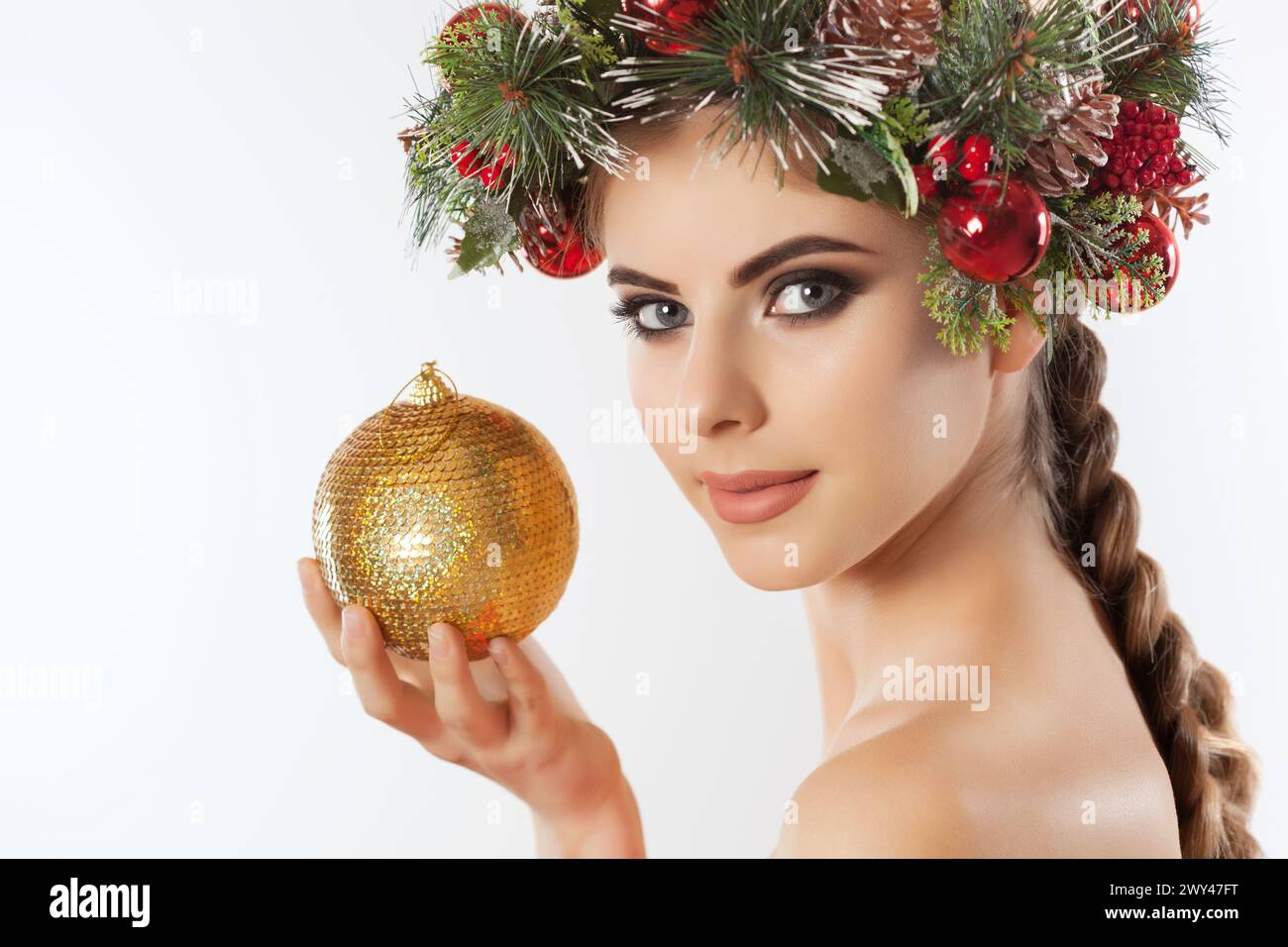 Une jolie jeune femme tient dans sa main une boule de Noël dorée, sur sa tête une belle couronne d'épicéa avec des cônes et des boules. Concept du nouvel an. Banque D'Images