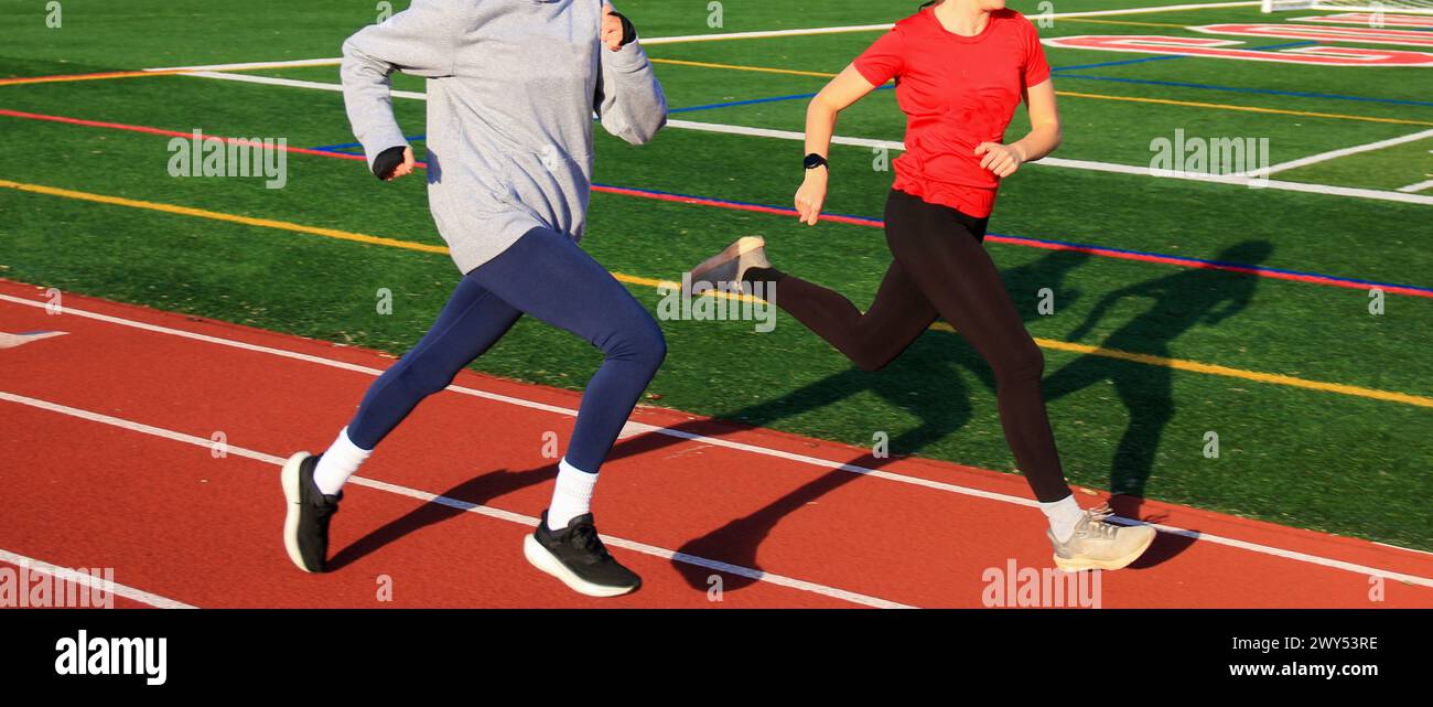 Deux lycéennes qui courent vite ensemble sur une piste pendant l'entraînement par une journée ensoleillée laissant des ombres sur le gazon. Banque D'Images