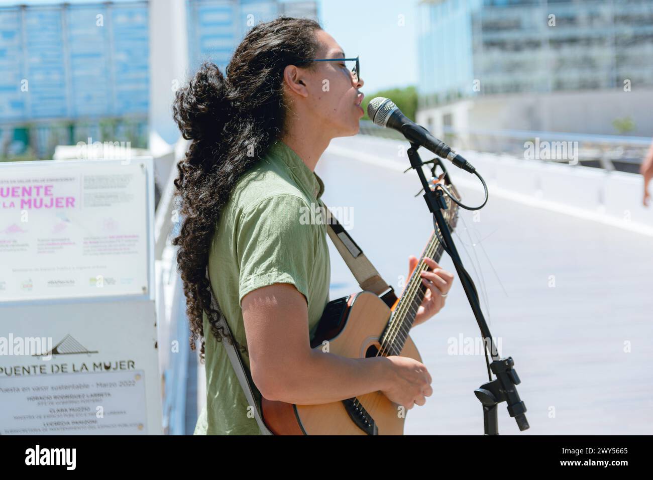 Vue de profil d'un jeune homme de busker avec de longs cheveux bouclés avec des lunettes de soleil, en Argentine jouant de la guitare et chantant travaillant à midi, espace de copie. Banque D'Images