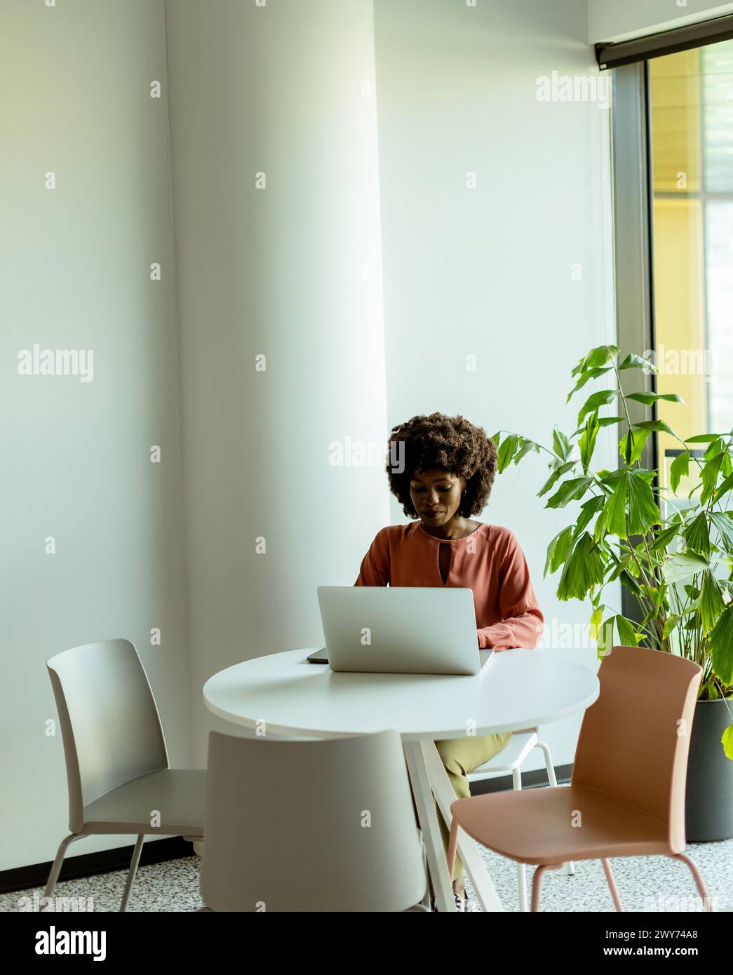 Une femme s'est concentrée sur son ordinateur portable à une table de café lumineuse près d'une grande fenêtre à côté de plantes d'intérieur. Banque D'Images