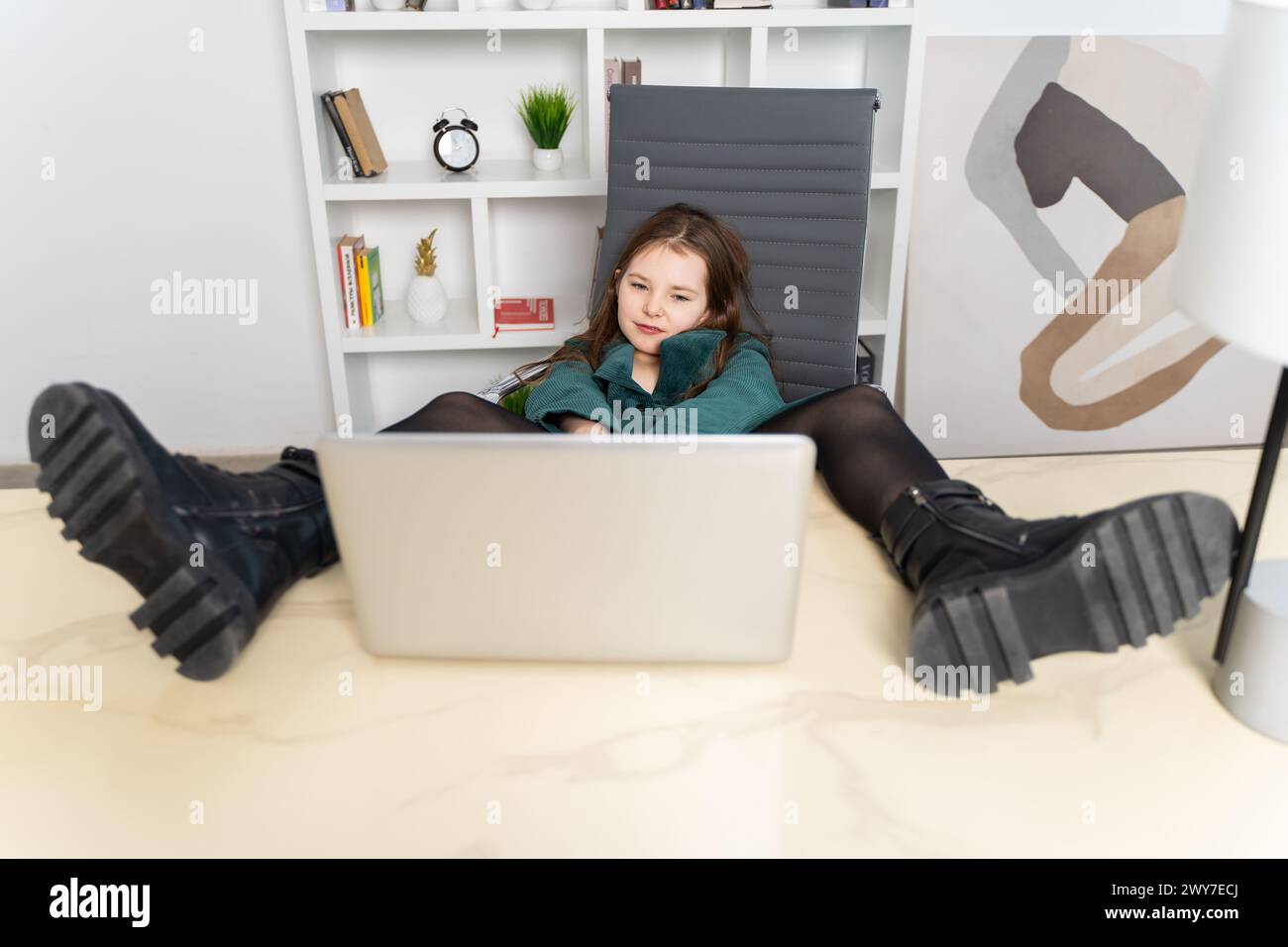 Petite fille assise dans une chaise de bureau avec ses jambes sur une table avec un ordinateur portable. Photo de haute qualité Banque D'Images