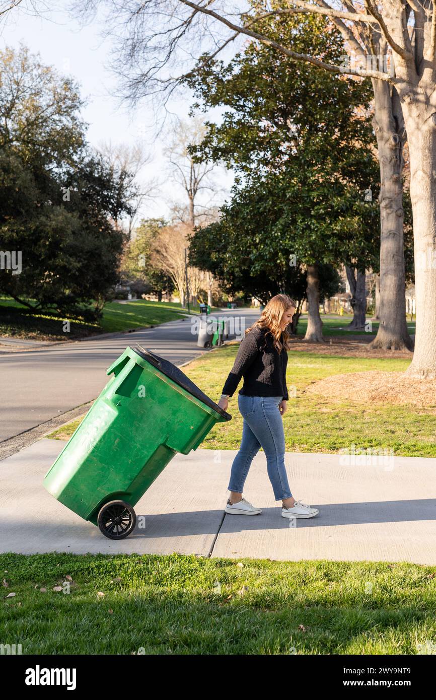 Femme roulant un bac de recyclage vert sur un trottoir. Banque D'Images