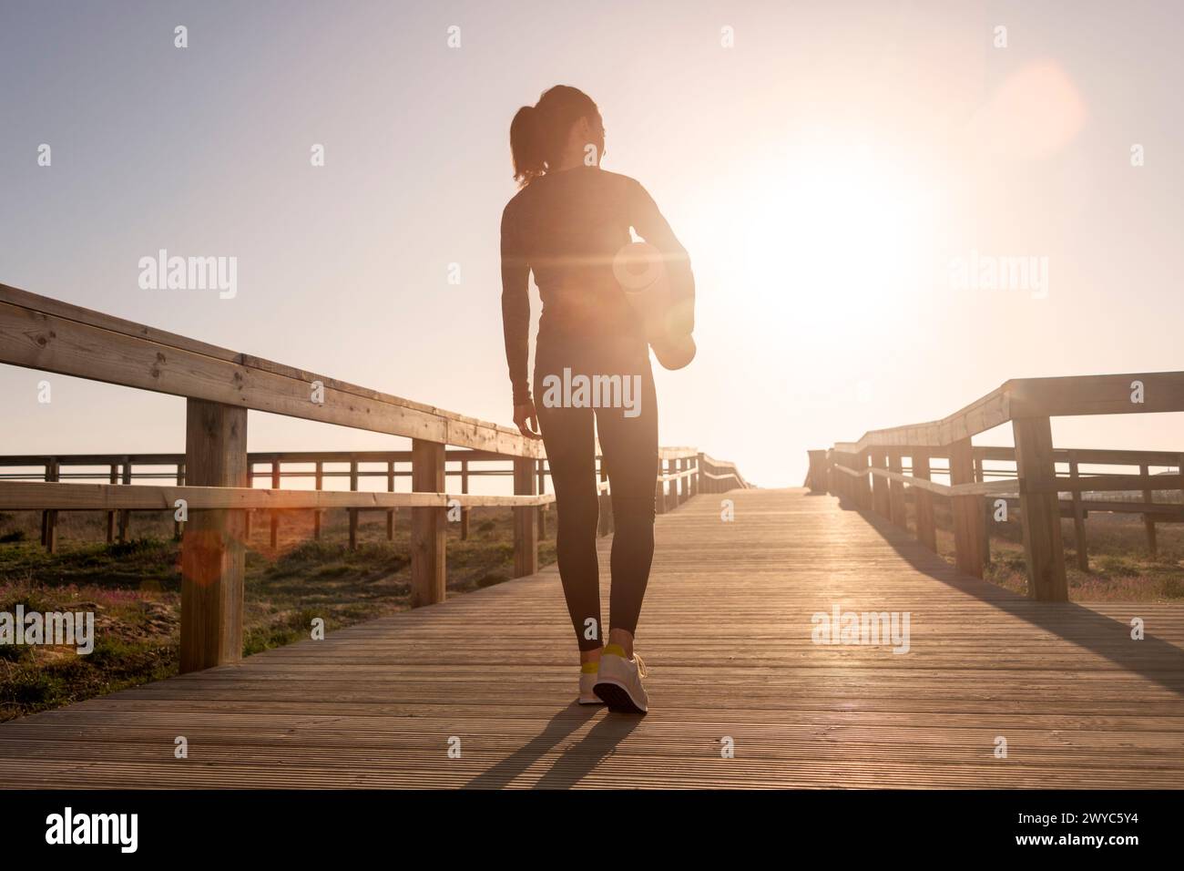 Vue arrière d'une femme sportive portant un tapis d'exercice, marchant le long d'une promenade au soleil Banque D'Images
