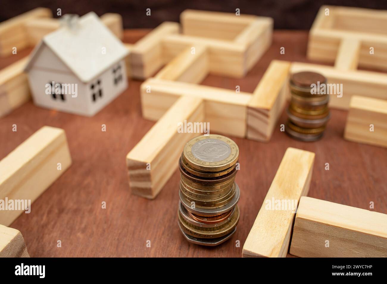 Piles de pièces de monnaie et une maison blanche miniature dans un labyrinthe en bois, métaphore des difficultés économiques Banque D'Images
