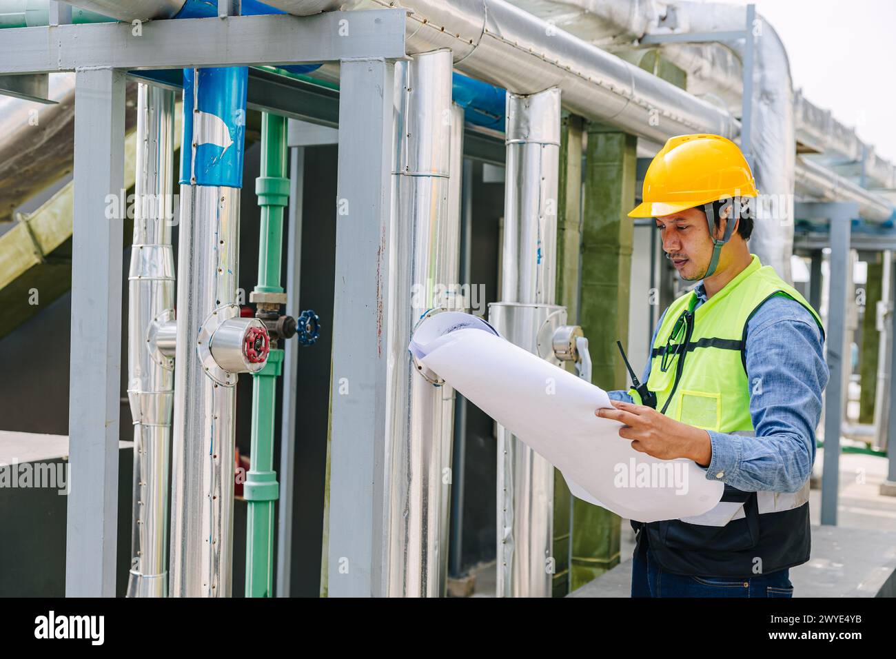 Un homme dans un casque jaune et un gilet vert regarde un morceau de papier. Il porte un casque de sécurité et il est travailleur Banque D'Images