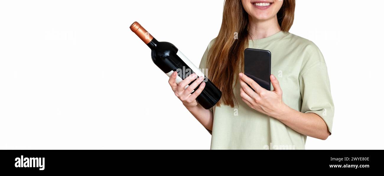 Jolie femme avec de longs cheveux bruns se tient devant un fond blanc tient une bouteille de vin et son téléphone portable dans ses mains et souriant. Banque D'Images