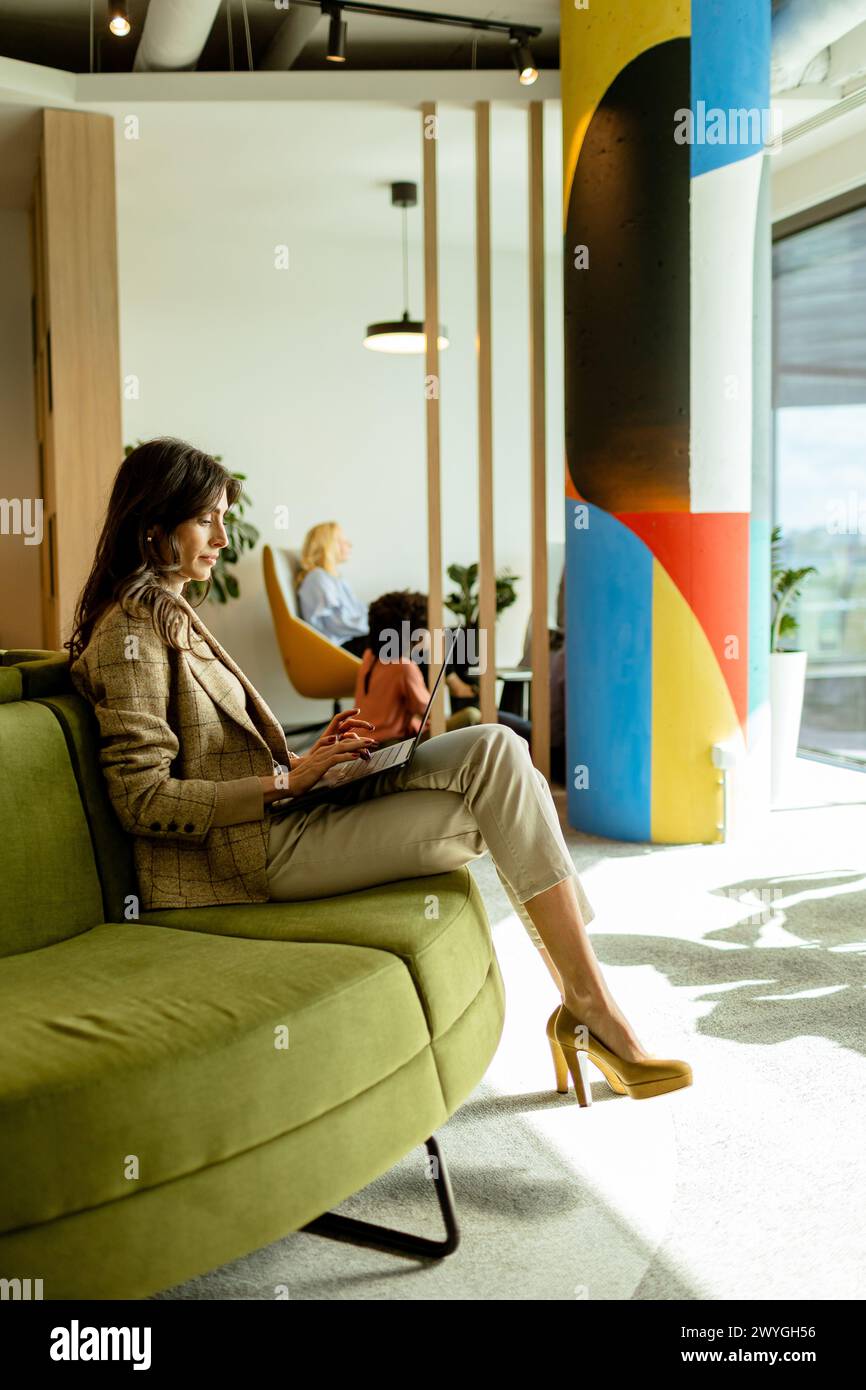 Focused Woman travaille sur son ordinateur portable, assise sur un canapé vert chic dans un bureau contemporain bien éclairé Banque D'Images