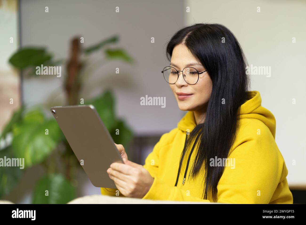 Une femme portant des lunettes regarde une tablette. Elle porte un sweat à capuche jaune. La tablette est un produit Apple Banque D'Images