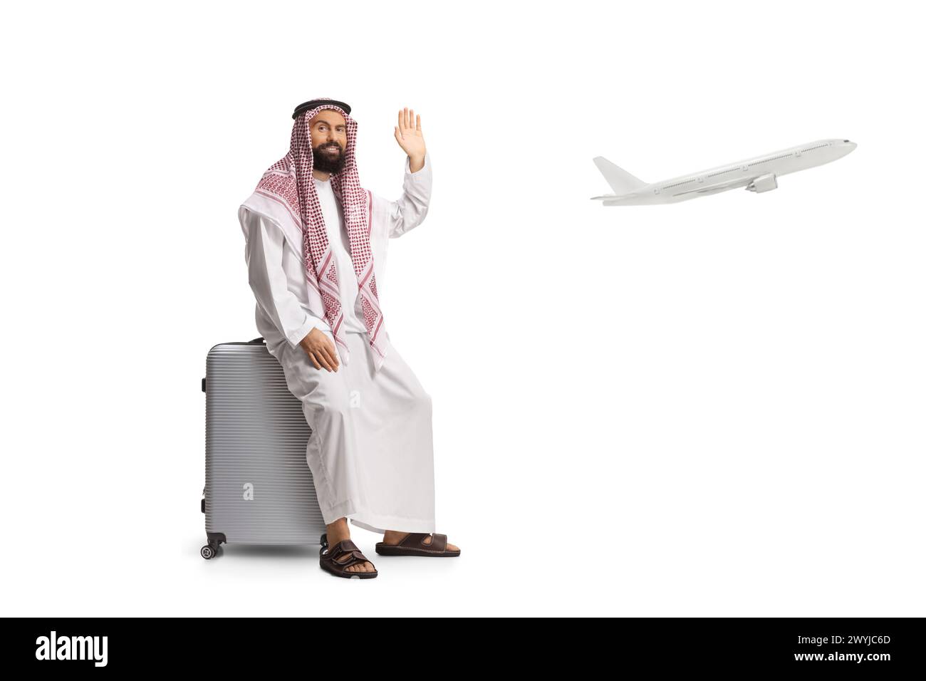 Homme arabe saoudien assis sur une valise à l'aéroport et agitant isolé sur fond blanc, avion volant dans le dos Banque D'Images