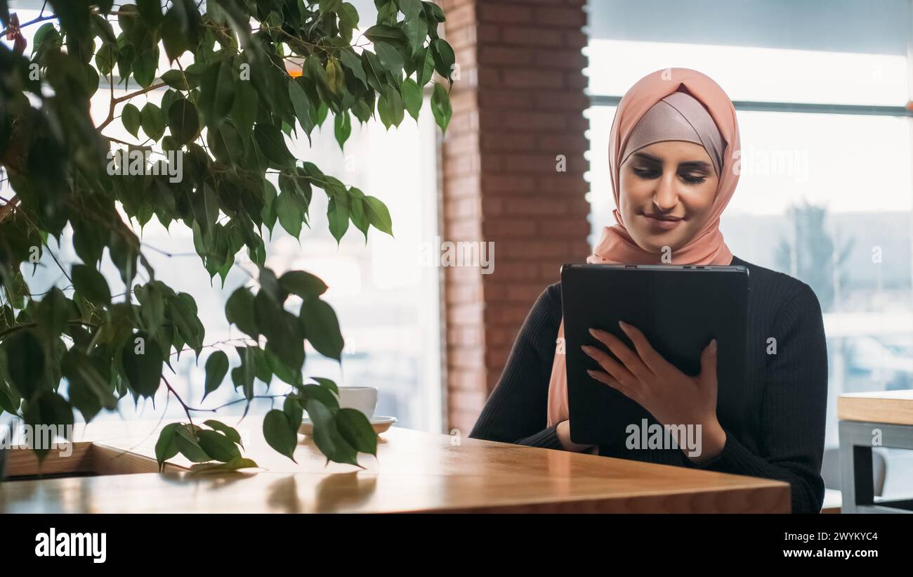 Travail en ligne. Tâche distante. Joyeuse femme freelance dans le hijab chat en ligne sur tablette numérique naviguant sur Internet au café confortable. Banque D'Images