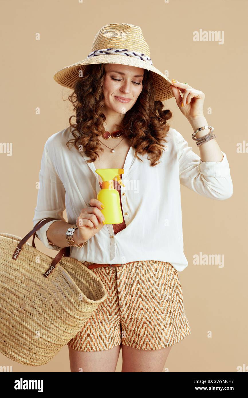 Vacances à la plage. femme tendance souriante en blouse blanche et short isolé sur beige avec sac de paille, spf et chapeau de paille. Banque D'Images