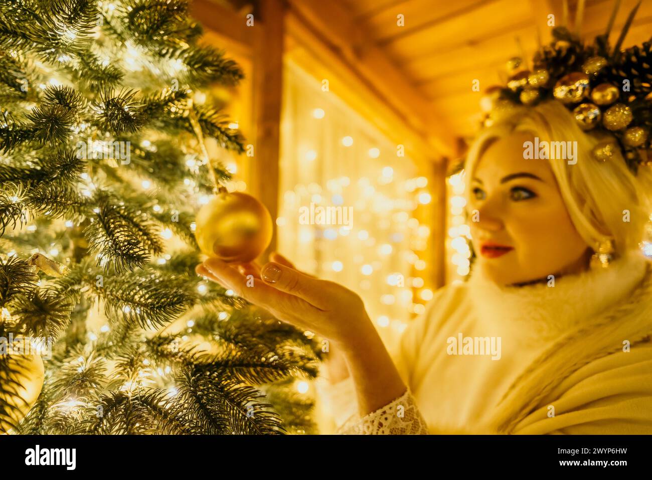 Une femme blonde en robe blanche et une couronne d'ornements en or décorent l'arbre de Noël avec des ornements en or et des lumières. L'arbre est décoré d'or Banque D'Images