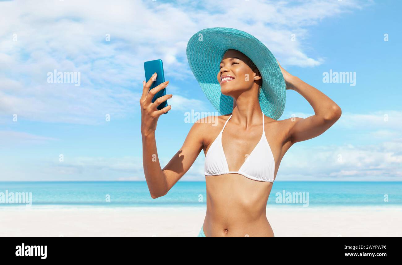 Heureuse jeune femme au bord de la plage à l'aide d'un téléphone portable, portant un chapeau de soleil turquoise et un bikini, portrait d'une femme africaine latino-américaine en summ ensoleillé Banque D'Images