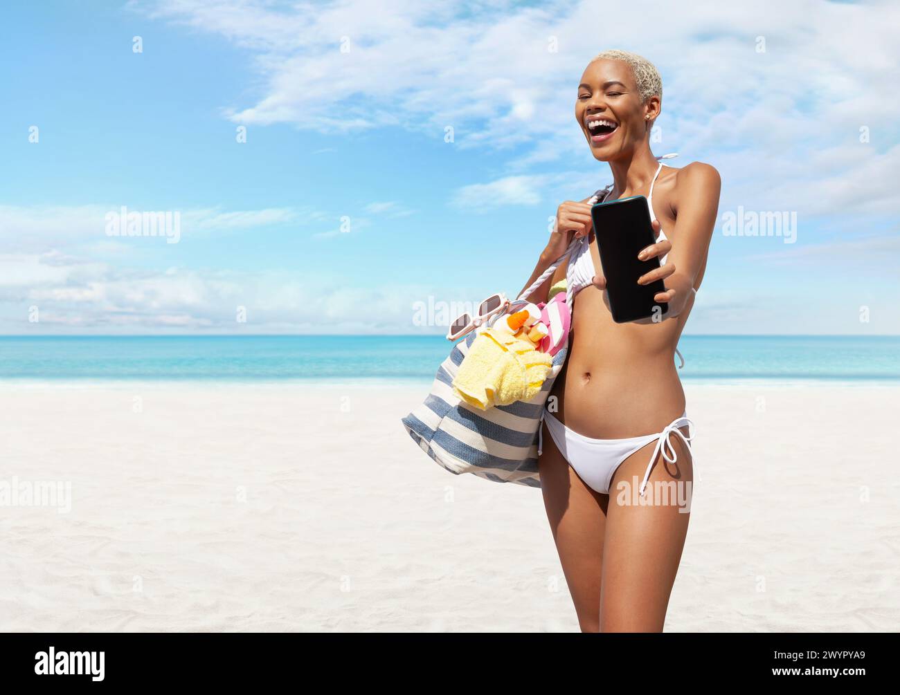 Femme heureuse au bord de la plage portant un bikini tenant un sac de plage et montrant un téléphone portable dans une journée ensoleillée avec un ciel bleu. Concept de vacances de plage d'été Banque D'Images