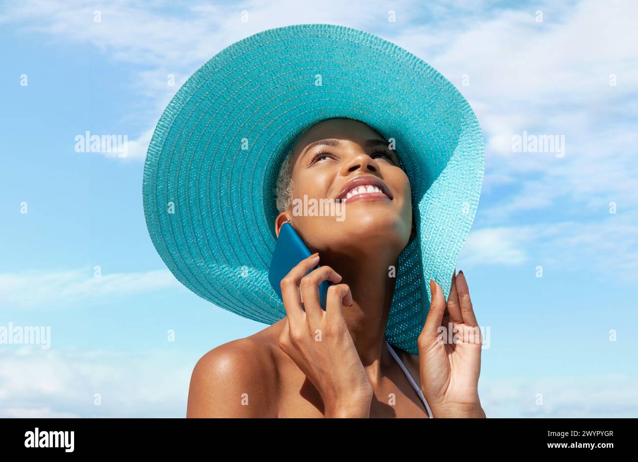 Femme heureuse au bord de la plage portant un chapeau de soleil bleu et utilisant un téléphone portable dans une journée ensoleillée avec un ciel bleu. Concept de vacances à la plage d'été, shopping onli Banque D'Images