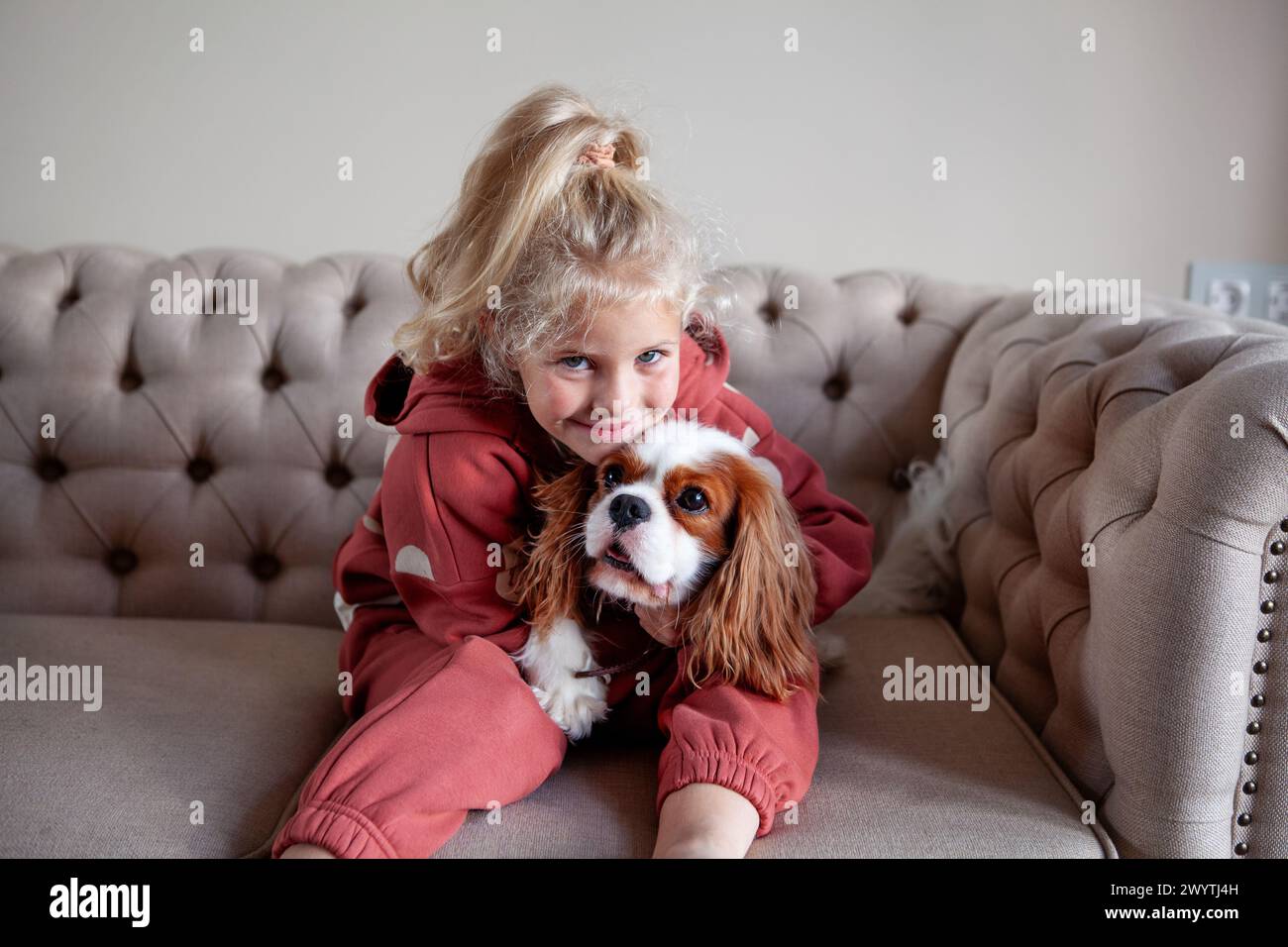 Belle fille blonde enfant est assis à la maison sur un canapé, jouant avec et embrassant le chien, créant des moments de joie et de compagnie avec l'animal de compagnie. Banque D'Images