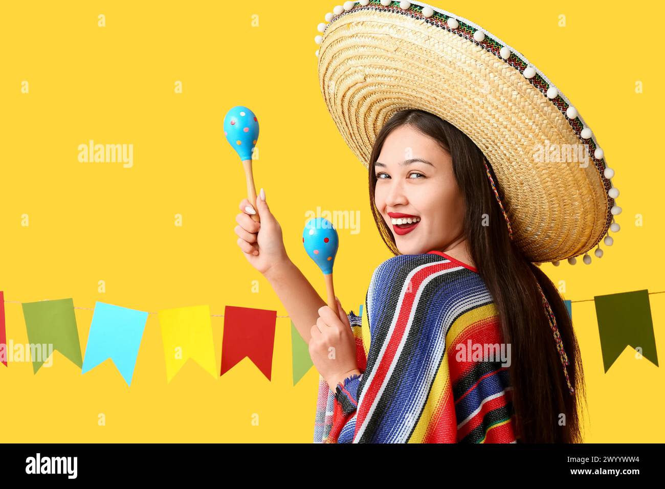 Heureuse jeune femme au chapeau sombrero mexicain et poncho tenant des maracas sur fond jaune Banque D'Images