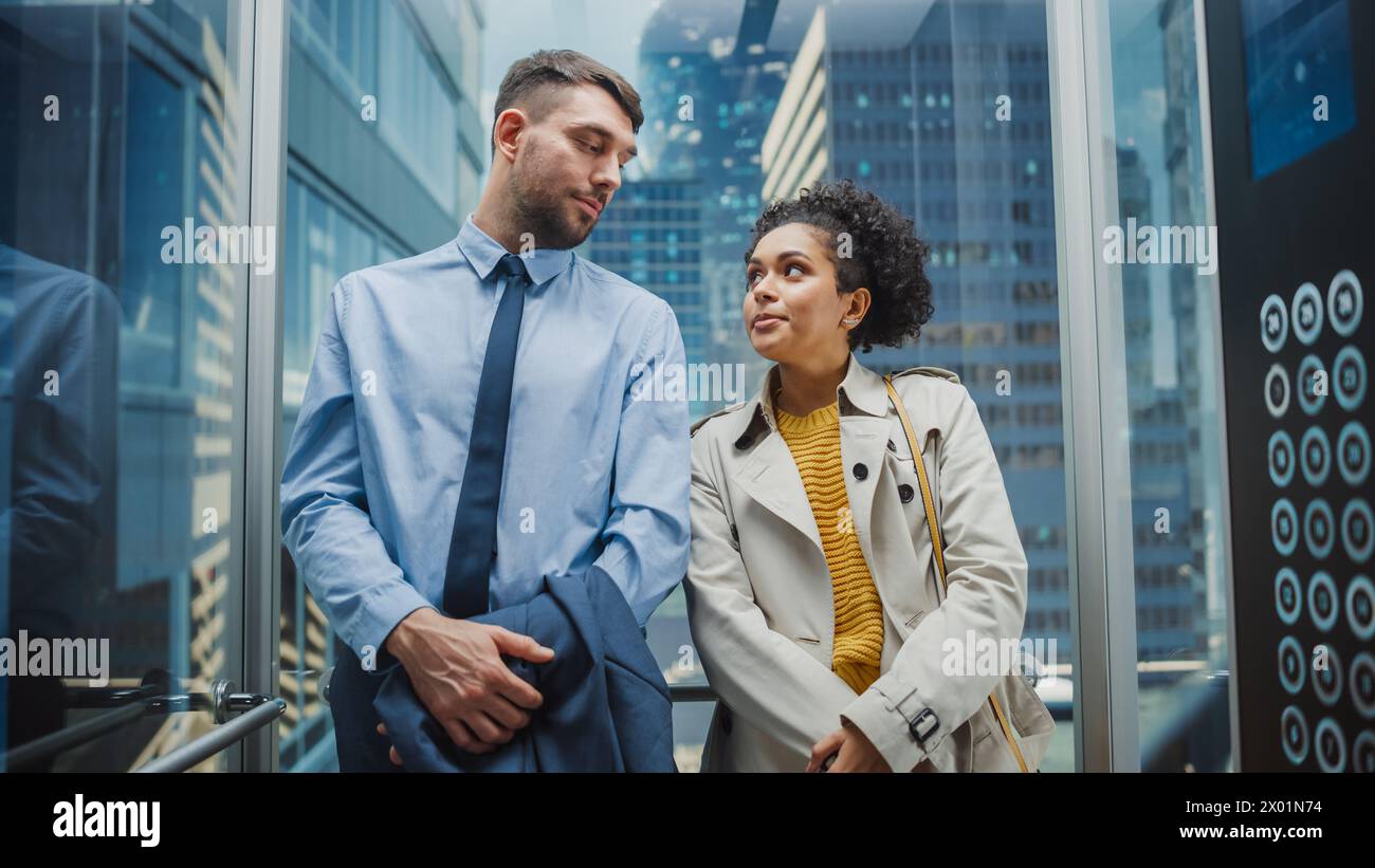 Deux collègues de bureau se regardant tout en roulant dans Glass Elevator dans un centre d'affaires moderne. Spécialiste caucasien masculin et responsable féminine noire d'Amérique latine discutez en toute décontraction dans l'ascenseur. Banque D'Images