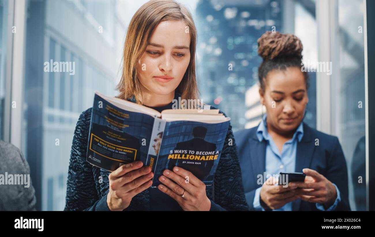 Confiant jeune femme Riding Glass ascenseur au bureau dans le centre d'affaires moderne. Manager junior lisant un livre financier intitulé « 5 façons de devenir millionnaire » dans un ascenseur. Banque D'Images