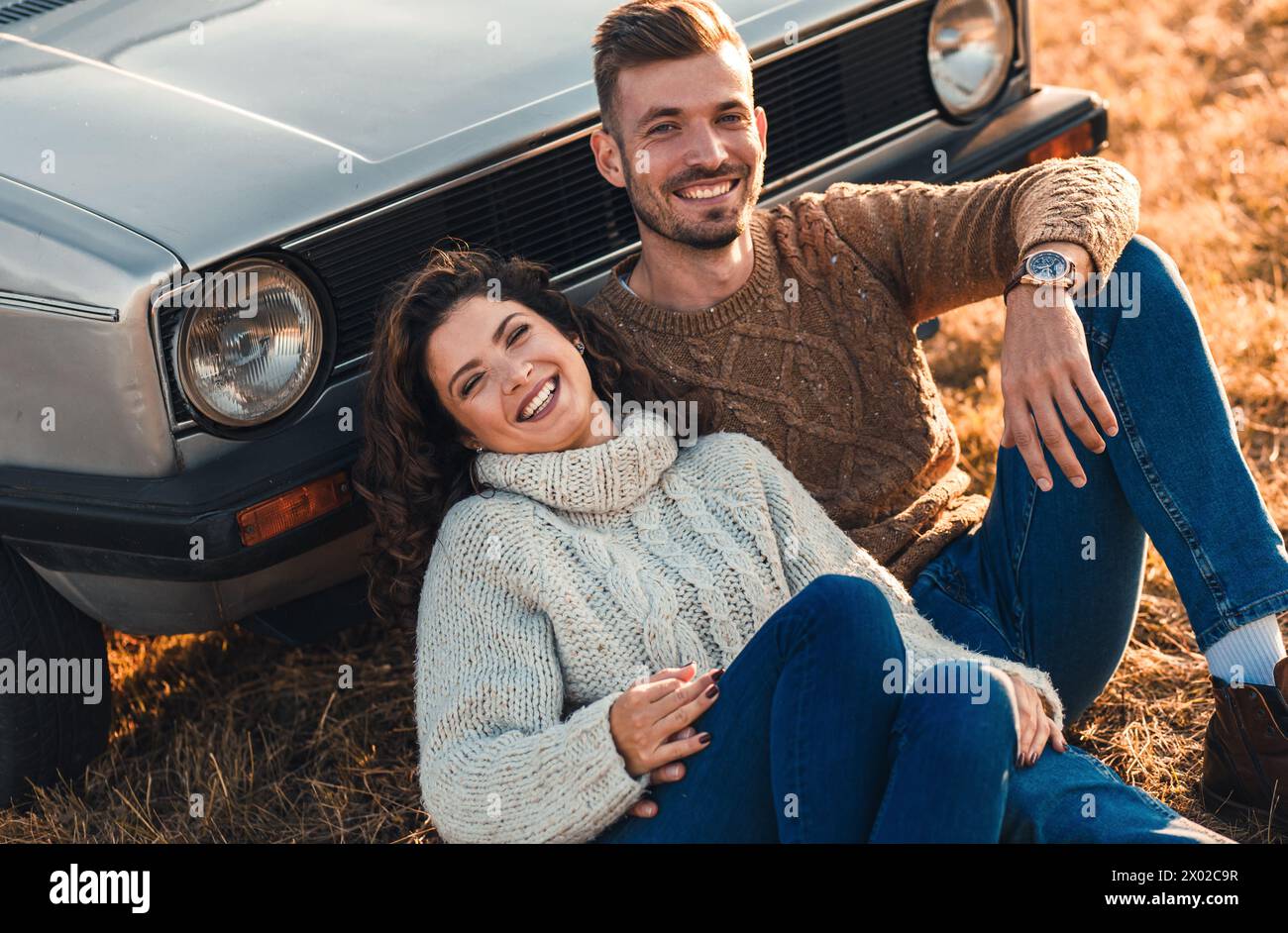 Beau jeune couple profitant du temps ensemble assis en plein air sur la prairie s'appuyant sur la vieille voiture embrassant les uns les autres. Banque D'Images