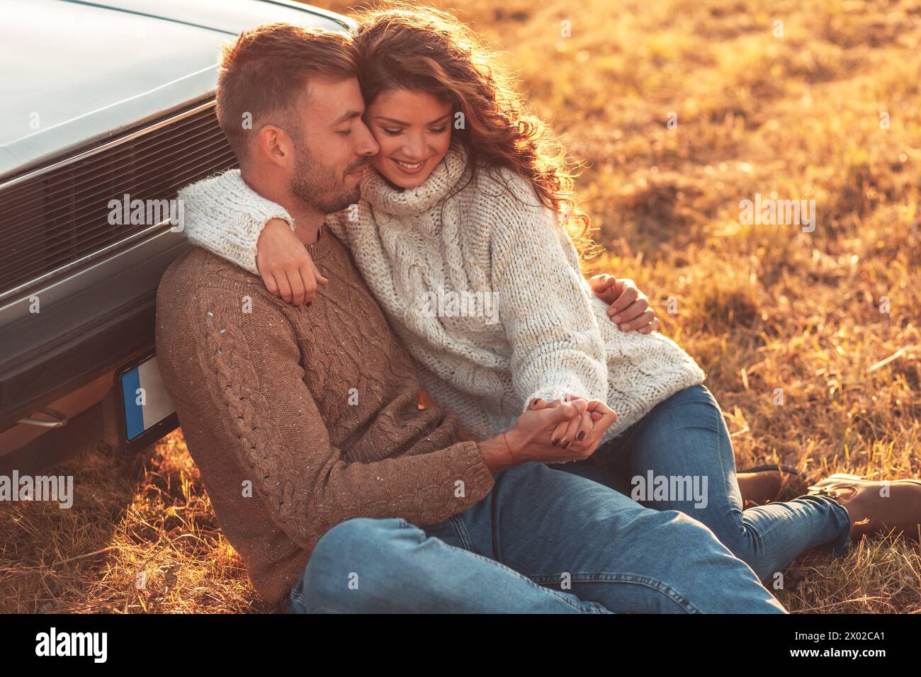 Beau jeune couple profitant du temps ensemble assis en plein air sur la prairie s'appuyant sur la vieille voiture embrassant les uns les autres. Banque D'Images